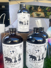 Bottles of Elderbeary® Syrup displayed on a wooden stand at an outdoor market, with a rustic background and product branding featuring a bear and forest theme.