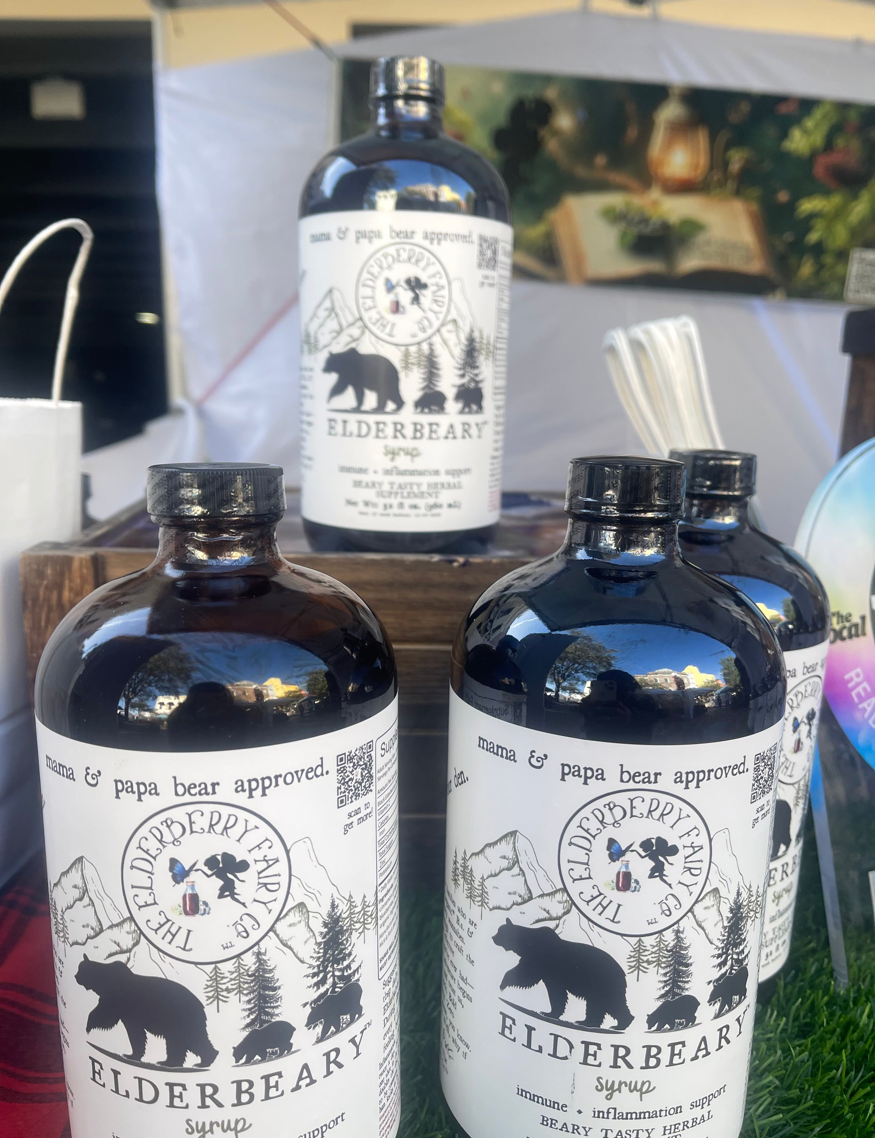 Bottles of Elderbeary® Syrup displayed on a wooden stand at an outdoor market, with a rustic background and product branding featuring a bear and forest theme.