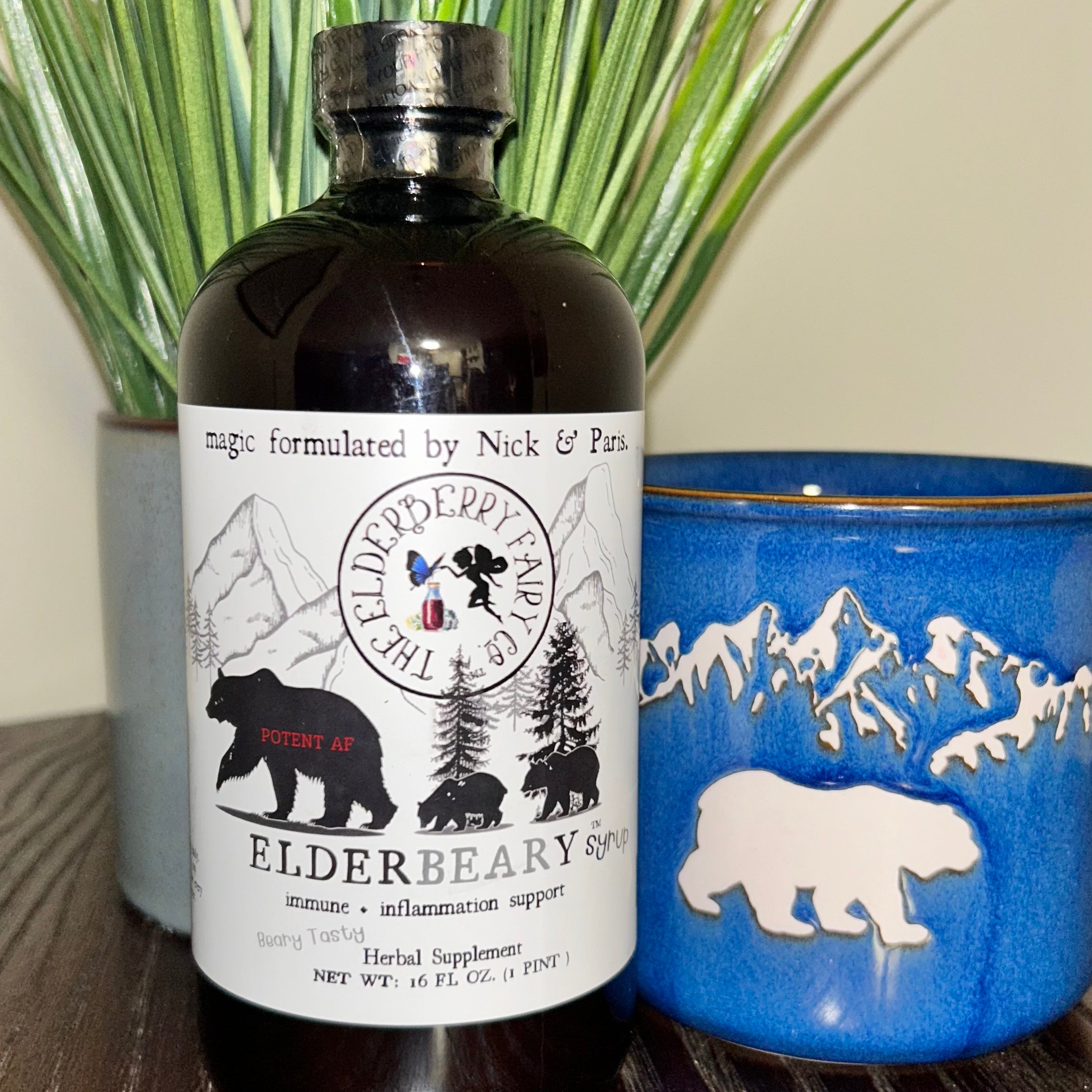 A bottle of Elderbeary® Syrup placed on a wooden surface, accompanied by green decorative grass and a blue ceramic mug with a white bear design.