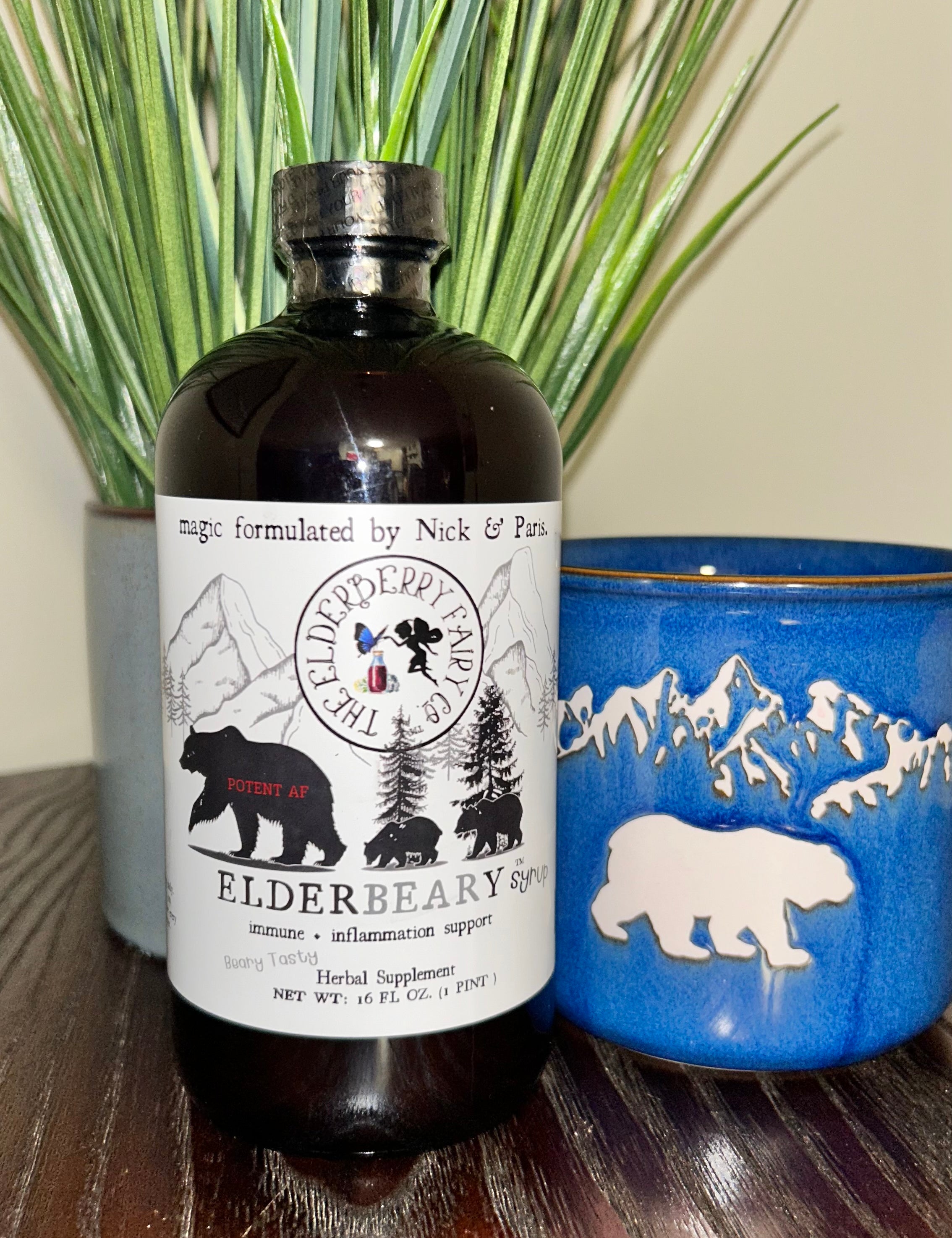 A bottle of Elderbeary® Syrup placed on a wooden surface, accompanied by green decorative grass and a blue ceramic mug with a white bear design.