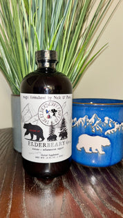 A bottle of Elderbeary® Syrup placed on a wooden surface, accompanied by green decorative grass and a blue ceramic mug with a white bear design.